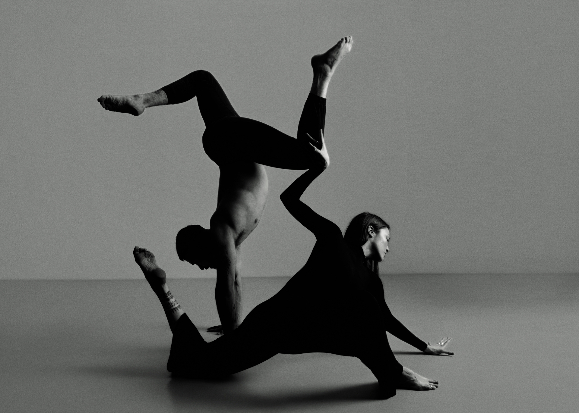 Two movement artists in a black-and-white studio composition — handstand and grounded balance